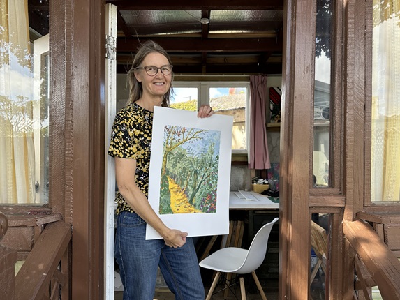 Wellington artist Carrie Carey, standing in a wooden doorframe, holding her A3 screen-print of a vibrant yellow pathway surrounded by autumn coloured bush. 