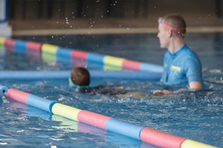 A child swimming down a pool lane with an instructor in blue standing next to them.