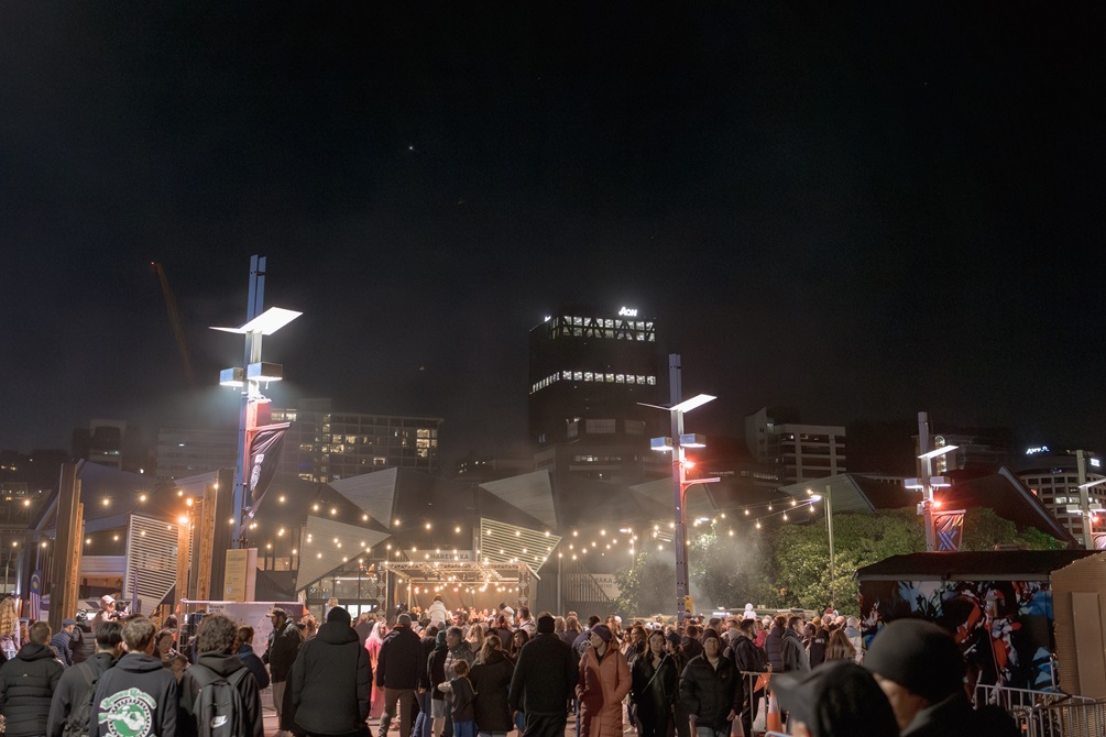 A thronging crowd of people and lights is contrasted against the dark sky above the Wellington building skyline.