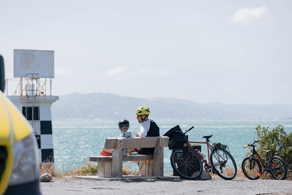 A father and son sit on a wooden park bench next to a small light house, looking out over the Wellington harbour.