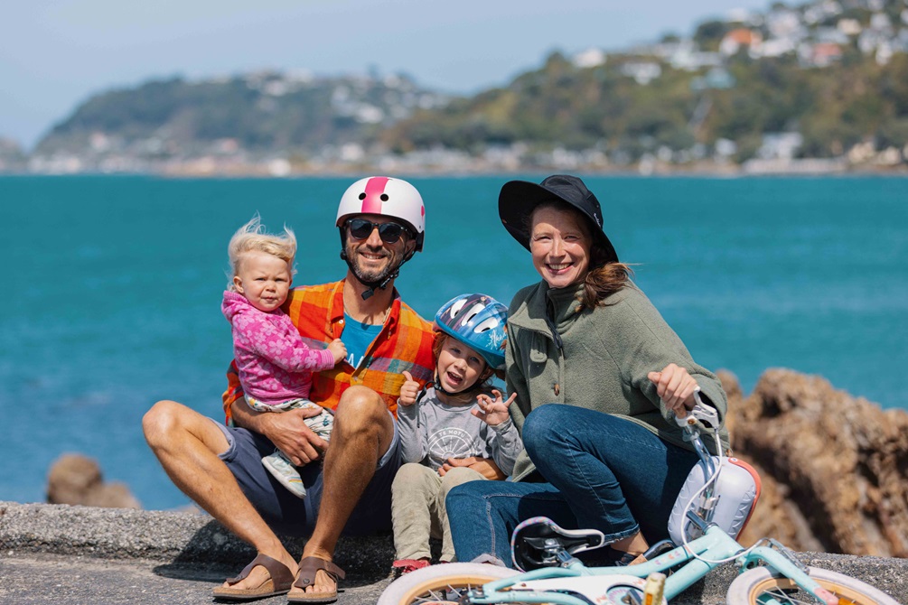 A family sit grinning on the side of Wellington harbour with helmets on and a kids bike lying in front of them.