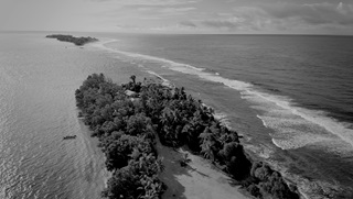 Black and white still from a drone video showing two small Pacific islands against a wide open sea and sky. In the foreground, the closest island is being beset by breaking waves.