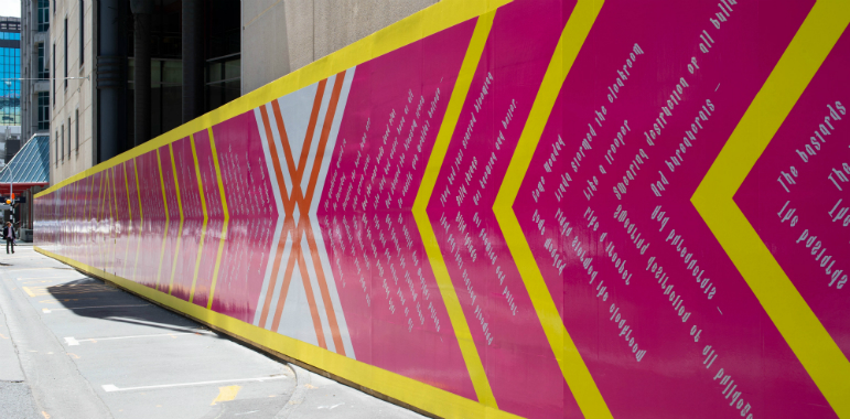 Civic precinct wall, decorated bright pink with bright yellow lines and white text accross it.