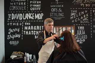 Woman having her hair cut in front of a blackboard listinghair-style options that have local politicians' names.