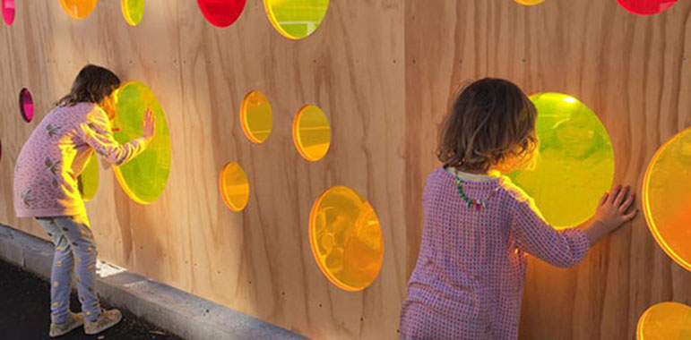 Children looking through colourful perspex 'windows' in plywood hoarding.