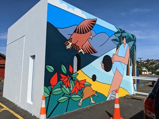 A mural painted on the side of a building, of a woman holding a ball and two native birds watching.