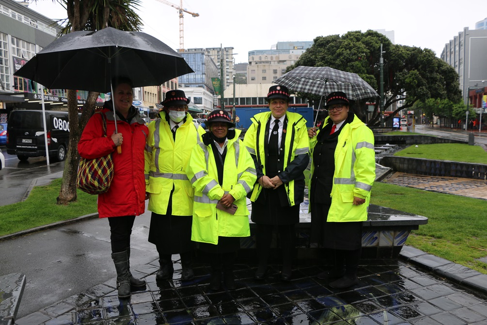 Four Māori wardens in high vis jackets with umbrellas standing in Te Aro park on a rainy grey Wellington day. They are joined by a woman in a red jacket too.