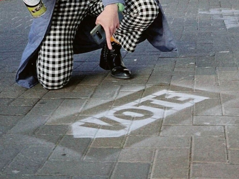 Someone kneeling and pointing to a Vote sign painted on the footpath.
