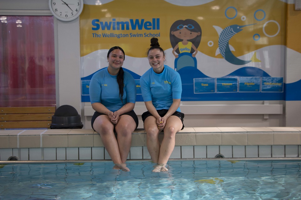 Aquatic Education Instructors Koopu Waipara and Milly Mackey, both 18, sit on the edge of the Tawa Pool smiling with their feet dangling in the water, with the colourful SwimWell sign behind.