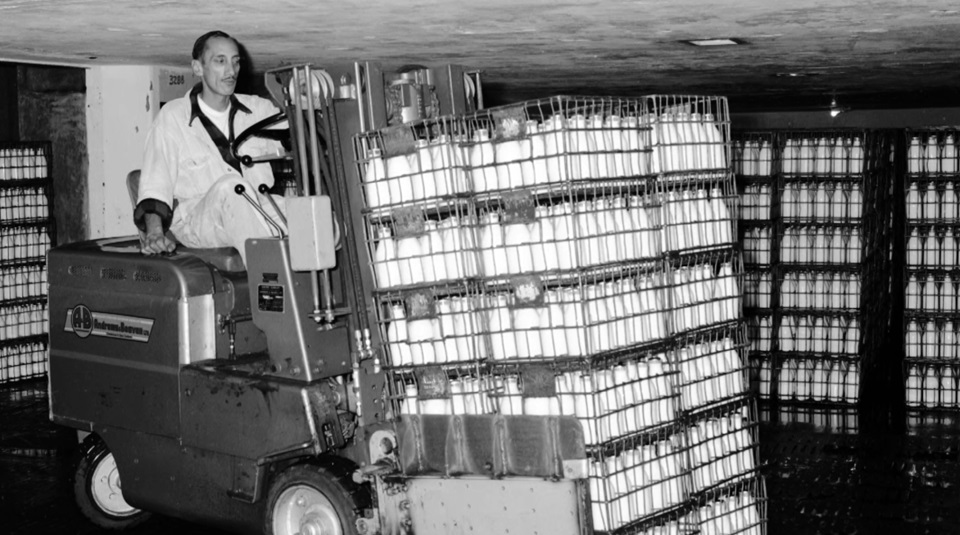 A man driving a forklift stacked with glass bottles filled with milk, surrounded by shelves also filled with milk bottles, at Wellington City Council's old Municipal Milk Department.