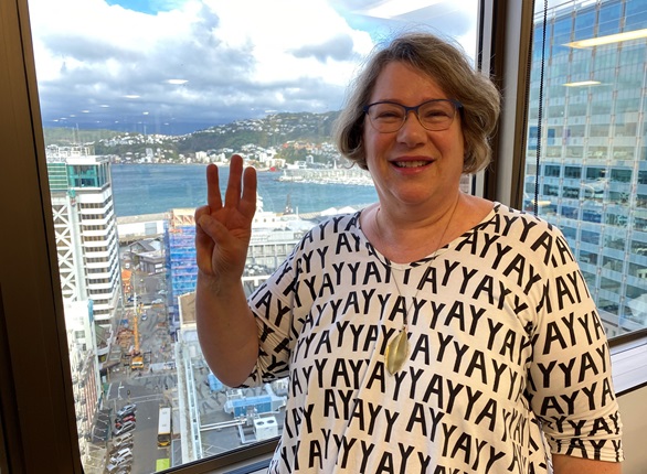 Rachel Noble in a white shirt with 'Yay' written all over it, smiling at the camera while doing sign language for 'Wellington', with the harbour in the background.