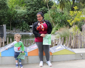 Bafrin, who is originally from Iran, wearing a bright pink top standing in front of the penguin enclosure at Wellington Zoo, smiling at a small boy looking down wearing a dinosaur jersey.