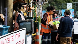 A staff member at the Tip Shop collecting a box of donations from a man and a woman, with a quirky sailor life-sized model as well as other random items in shot.
