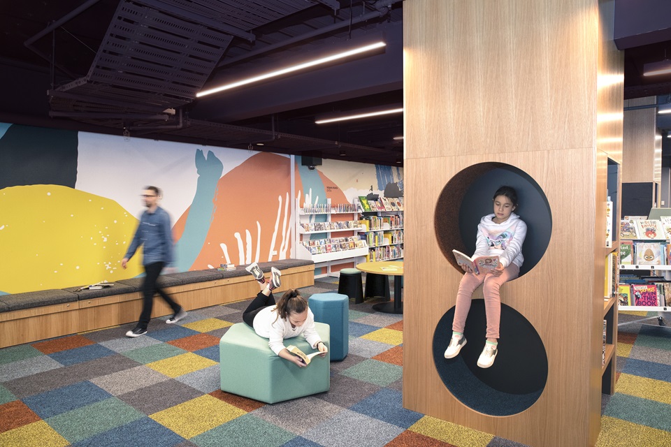 A girl reading a book sitting up high in a circular cubby hole at Te Awe Library, with a man walking past and another girl reading on a comfy, wide rectangular seat.