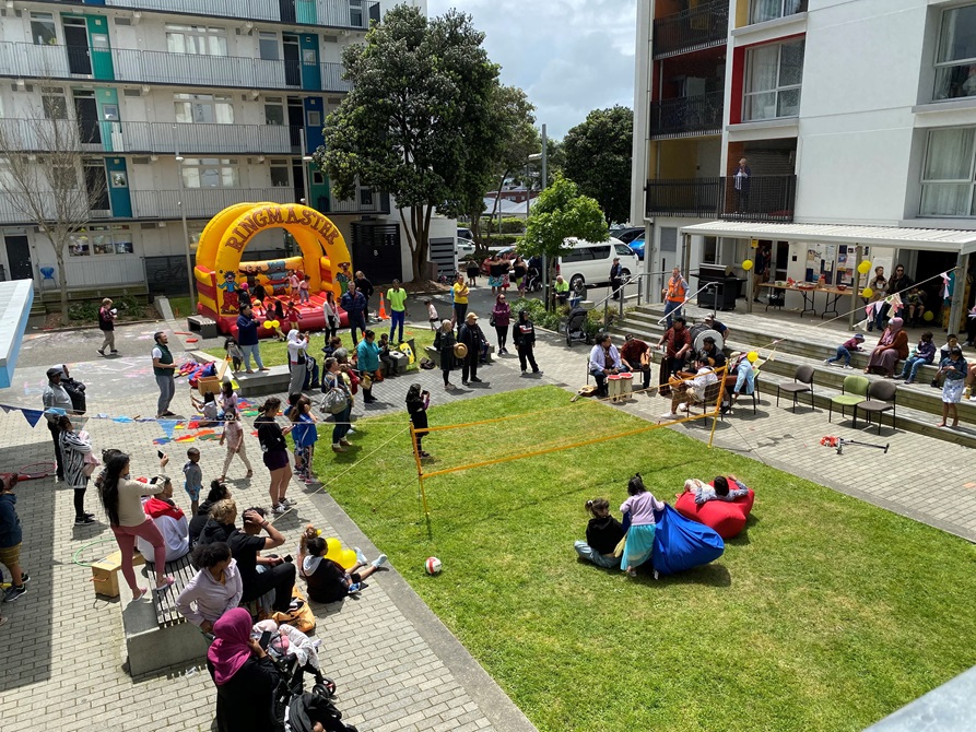 A high angle shot of the Newtown Apartment complex on a sunny day with families outside playing volleyball and on a bouncy castle, sitting on the grass, and enjoying music and dance.