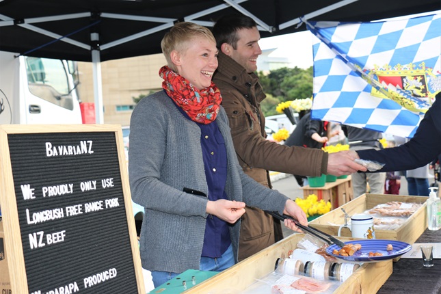 Lena Donandt with a big smile with a blue plate of cut up sausages, and Sebastian Nebel handing over some of his handmade German-style sausages to a customer, with a Bavarian flag hanging on the side of their marquee stall.