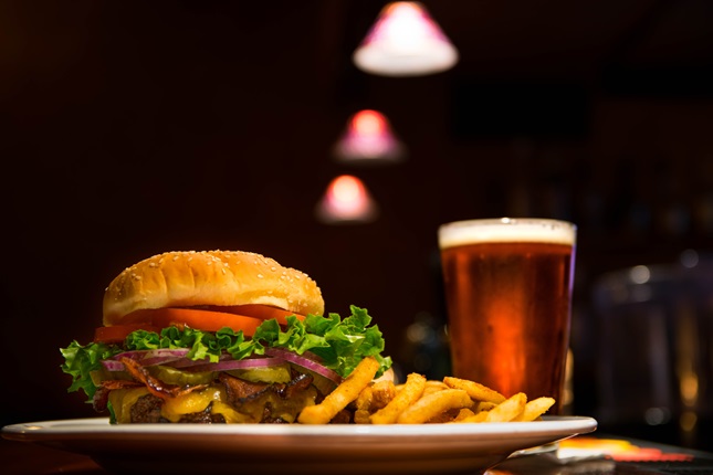 A close-up shot of a plate with a burger and fries on it, and to the left, a beer, with a dark background and blurred lights above.