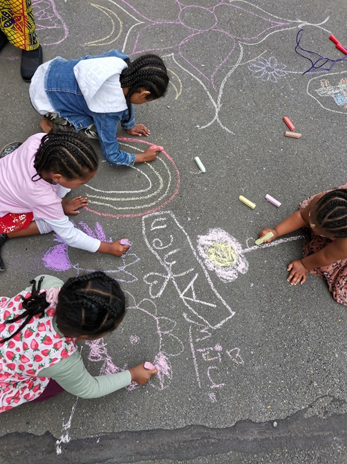 An overhead shot of children with braids and ponytails drawing shapes and rainbows in colourful chalk on the concrete. 