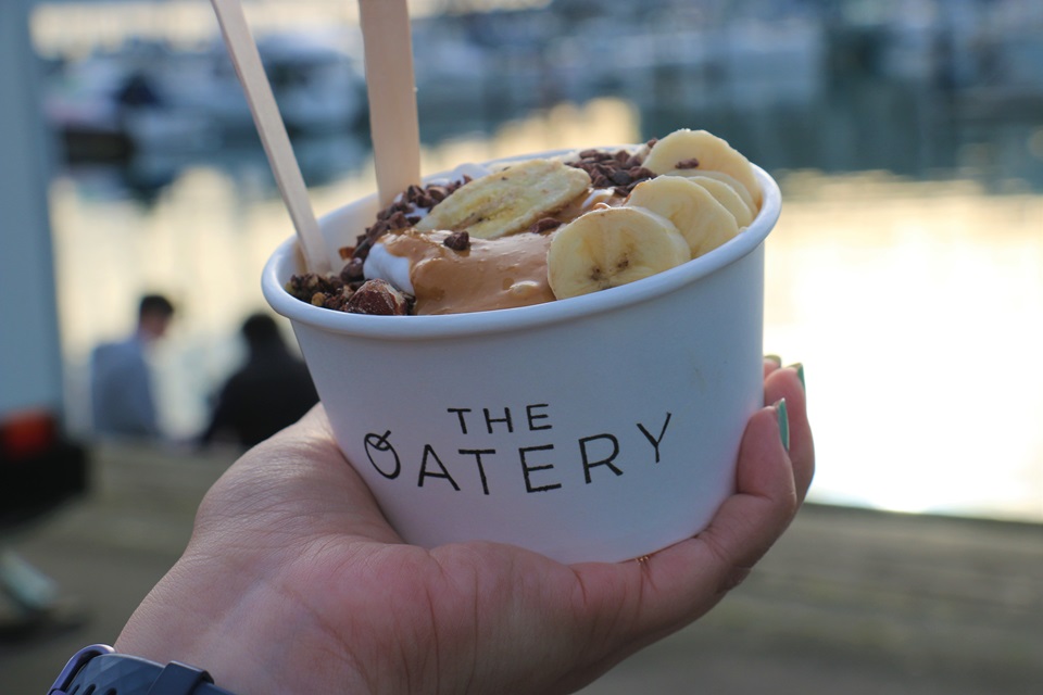 A hand holding a white paper cup with the words The Oatery in black, filled with oats, banana, and peanut butter toppings, with the harbour out of focus in background.
