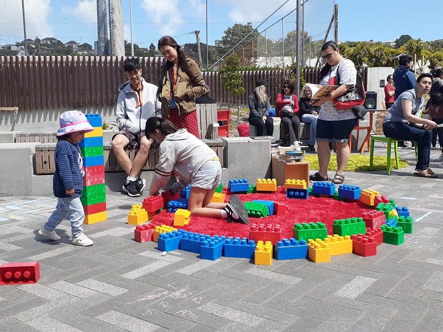 Children playing with a Lego circle outdoors on the concrete, with parents supervising from a wooden bench.