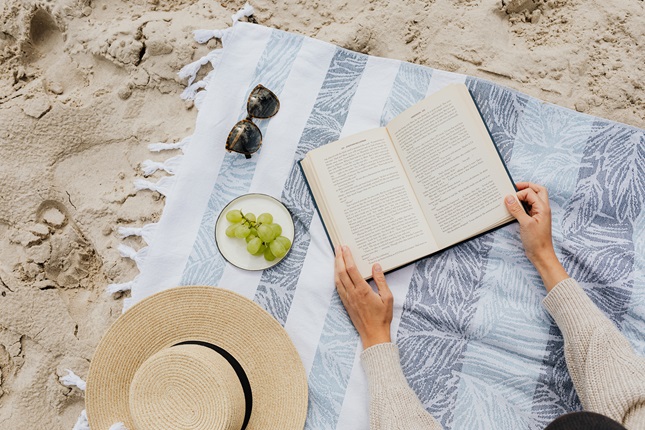 A birds-eye view of a blue and white striped towel on the sand, with a hat, some sunglasses, a drink, and a book.