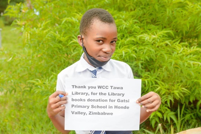 A primary school pupil from Zimbabwe, standing in greenery, holding a sign that thanks Council's Wellington City Libraries team for the books they sent to the school.