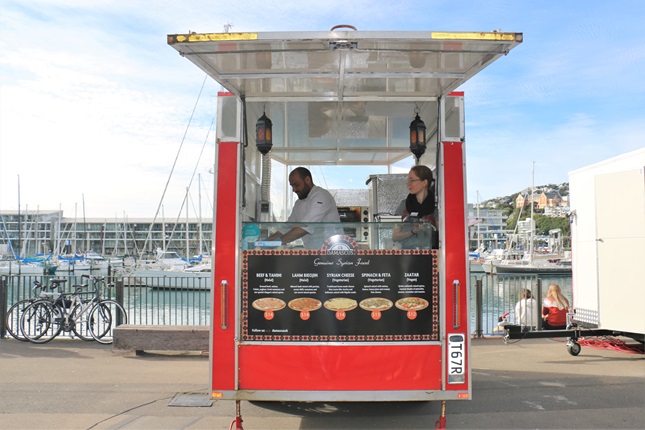 Hasan Alwarhani in his small, red food trailer, Damascus, which sells Syrian food, situated on Wellington Waterfront with the harbour in the background.