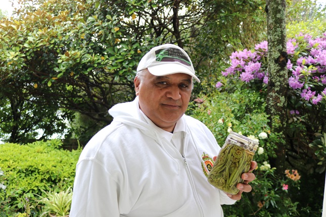 Chef and kaumatua Joe McLeod, wearing a white cap and jersey, holding up a jar of pickled native New Zealand pikopiko, the young curled shoots of ferns, standing in his sunny backyard surrounded by bush and purple flowers.