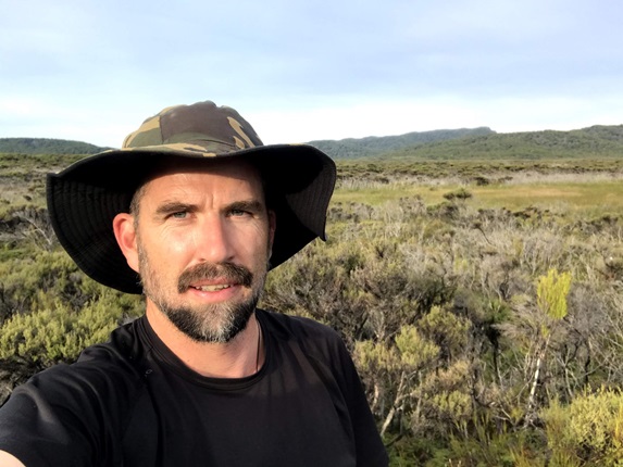 Tim Park pictured in a black t-shirt and wearing a camo hat with vast flat bush behind him, on Rakiura Stewart Island while on a trapping trip.