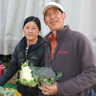  Fresh produce growers Queenie Jung on the left, and husband Tony on the right holding a cauliflower and a broccoli, smiling.