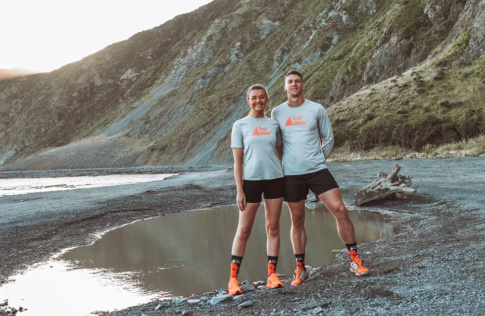 Frieda and Hardus de Bruyn, wearing matching grey t-shirts, black shorts, and bright orange running shoes, standing together on the South Coast beach with Red Rocks behind them.