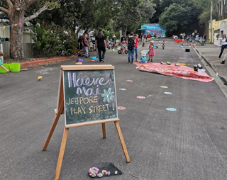 A chalkboard with the words 'haere mai', welcoming people to the Berhampore Play Day event, with lots of kids and adults playing on the street behind the sign.