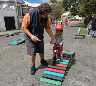 A man helping a young girl on her tricycle, navigating down a small colourful ramp, set up on a street closed to vehicles for a public play day.