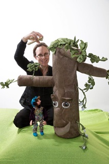 A woman holding a puppet in front of a model tree stump with a face.