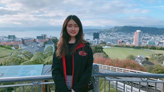 April Che, Cable Car Sales & Marketing Executive, standing on the Cable Car platform at the Botanic Garden with Wellington city and harbour in background.