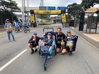 Four members from Dsport, one boy in a sports wheelchair and a man and two other children all smiling and crouching around him, on the road near Wellington city Lagoon at the start line of the Round the Bays fun run.