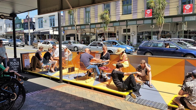 The bright yellow parklet located next to the pavement in Newtown, filled with 10 people at tables and sitting on the platform playing backgammon.
