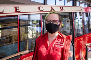 Jessica Eden, Cable Car Passenger Service Operative and Administration, standing in her red uniform with a black mask over her mouth and nose, next to the iconic red Cable Car.