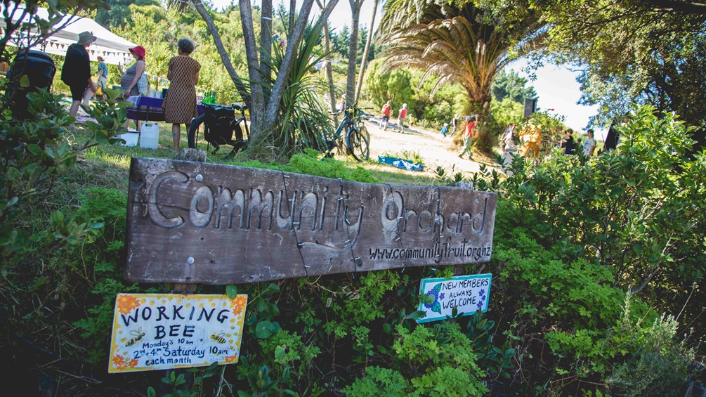 A wooden Community Orchard sign, with the gardens and people talking in the sun among trees in the background.