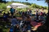 Lots of people sitting in the shade on picnic blankets, with a marquee and an orchard of trees in the sunny background.