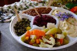 A round platter with various dips, including hummus, relish, and beetroot, with olives, vegetables, and purple flowers.
