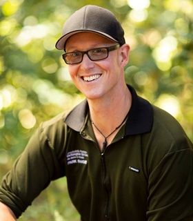A portrait of Wellington City Council Park Ranger Adam, sitting with a cap, his green uniform t-shirt, and blurred green leaves behind him.
