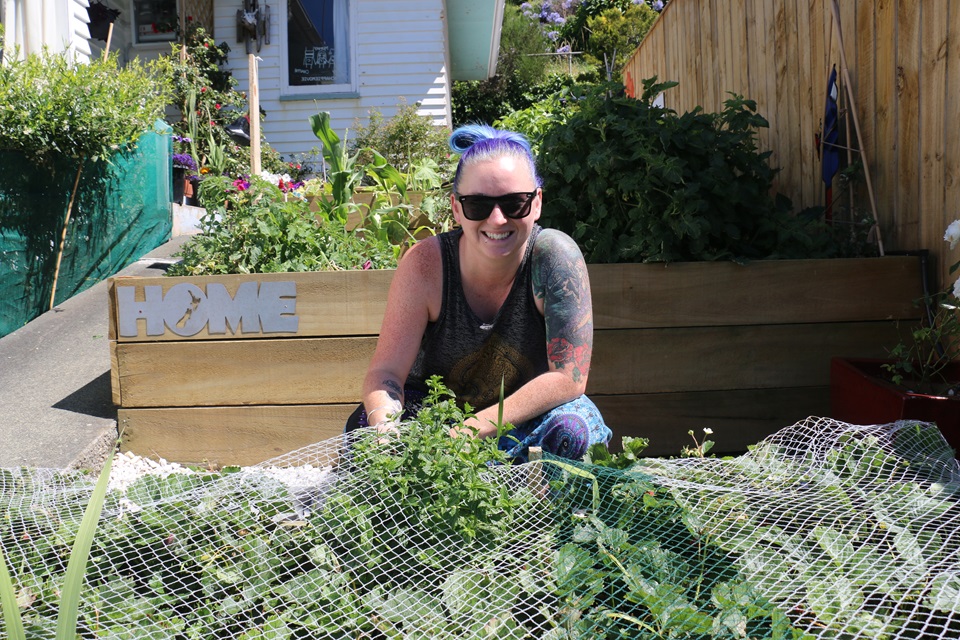 Miramar resident Ashley, wearing sunglasses, crouching behind her vegetable garden with another planter box and a white house behind her.