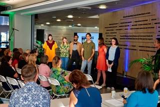 The six people who came up with the winning 'Ingrain' vision of transforming the food system by changing from annual to perennial crops, standing in a row in brightly coloured clothes, in front of an audience and the Wellington Climathon judging panel.