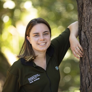 Wellington City Council park Ranger, Katie, in her green ranger uniform t-shirt, leaning her right arm up against a tree trunk, with dappled green leaves behind.