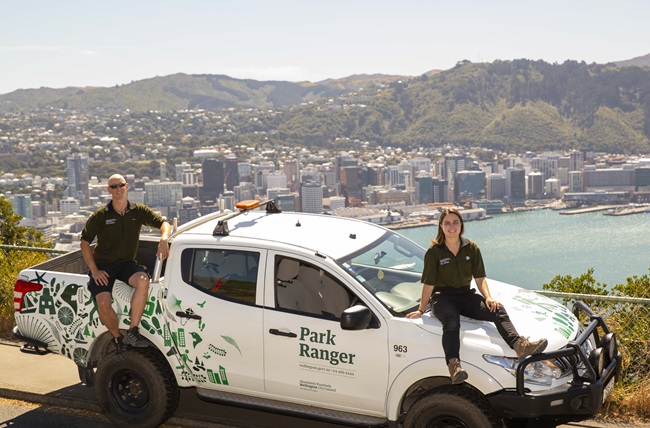 Two Wellington park Rangers, Adam on the left and Katie sitting on the right of a white ranger ute, parked up at Mount Victoria with a view of Wellington harbour in background.