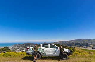 A bright blue sky with Wellington hills beyond, and a white ute parked on grass, with two park rangers, Adam on the left, Katie on the right, standing on either side of the work vehicle.