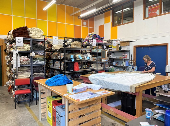 A Volunteer at the Curtain Bank cutting a large piece of white fabric on a workbench in the middle of a room, with shelves of stacked curtains and bright yellow and orange tiles on the wall.