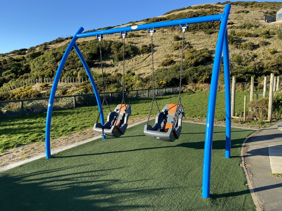 Accessible swings hanging off their blue frame at Tawhai Raunui Play Area, will green hills and blue sky in background.