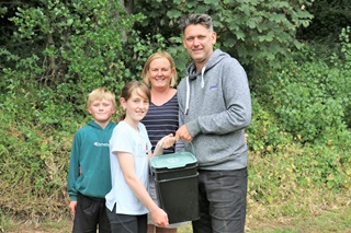 A young boy in a green sweatshirt, next to his sister in a white t-shirt, and their parents huddled together holding a black Bokashi bin with green lid, standing in front of green bush and trees.